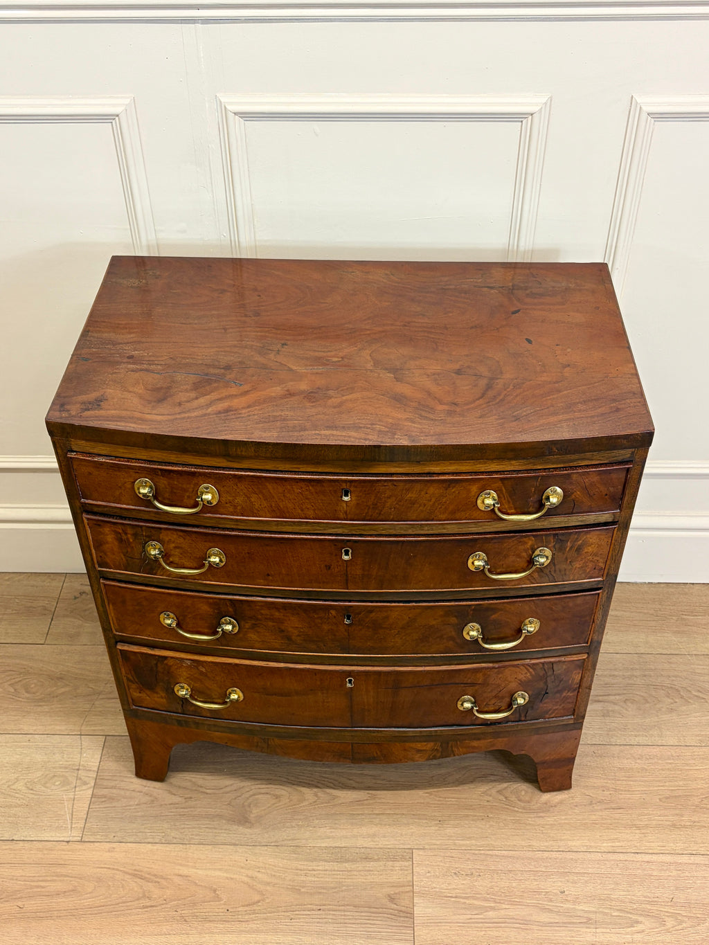 Wooden dresser with curved design and brass handles against a white paneled wall.