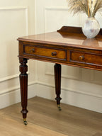 Wooden desk with a vase of flowers on a wooden floor against a white paneled wall.