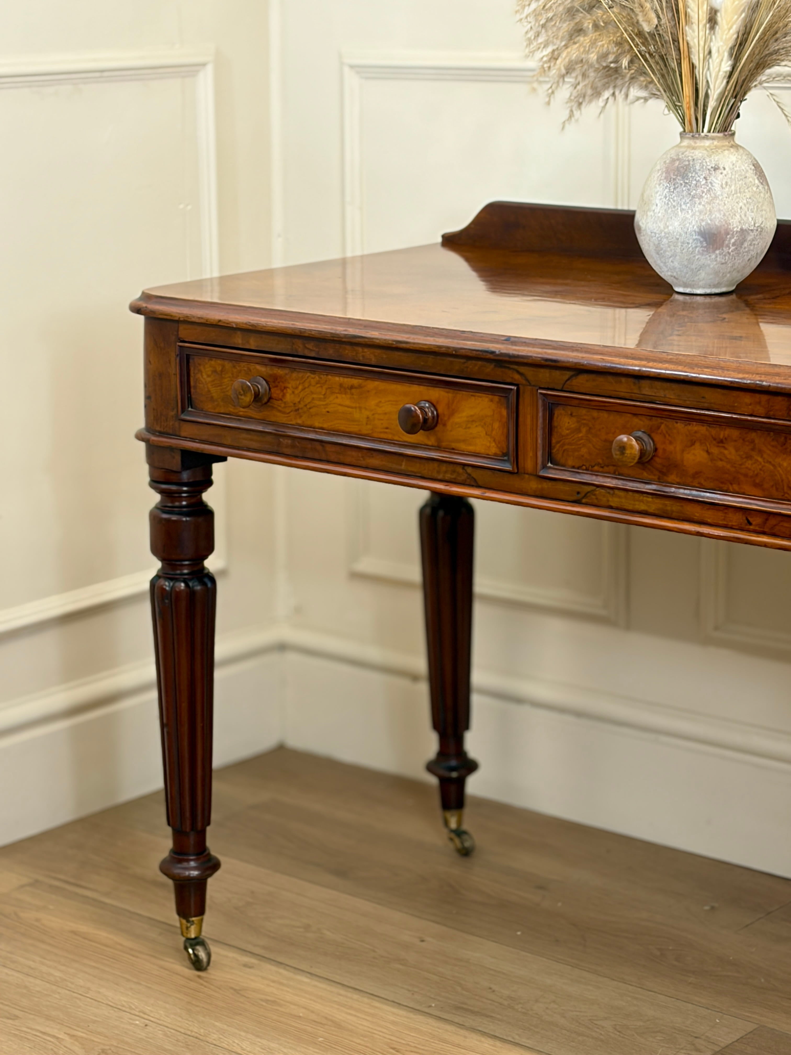 Wooden desk with a vase of flowers on a wooden floor against a white paneled wall.