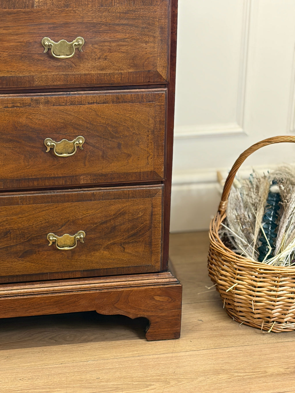 Wooden dresser with brass handles next to a wicker basket on a wooden floor.