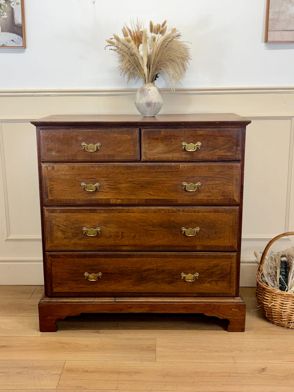 Wooden dresser with four drawers on a wooden floor, with a vase of dried plants on top.