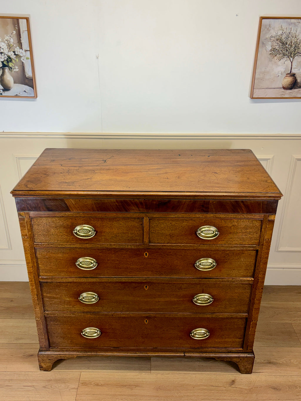 Wooden dresser with brass handles on a wooden floor with white walls and framed pictures.