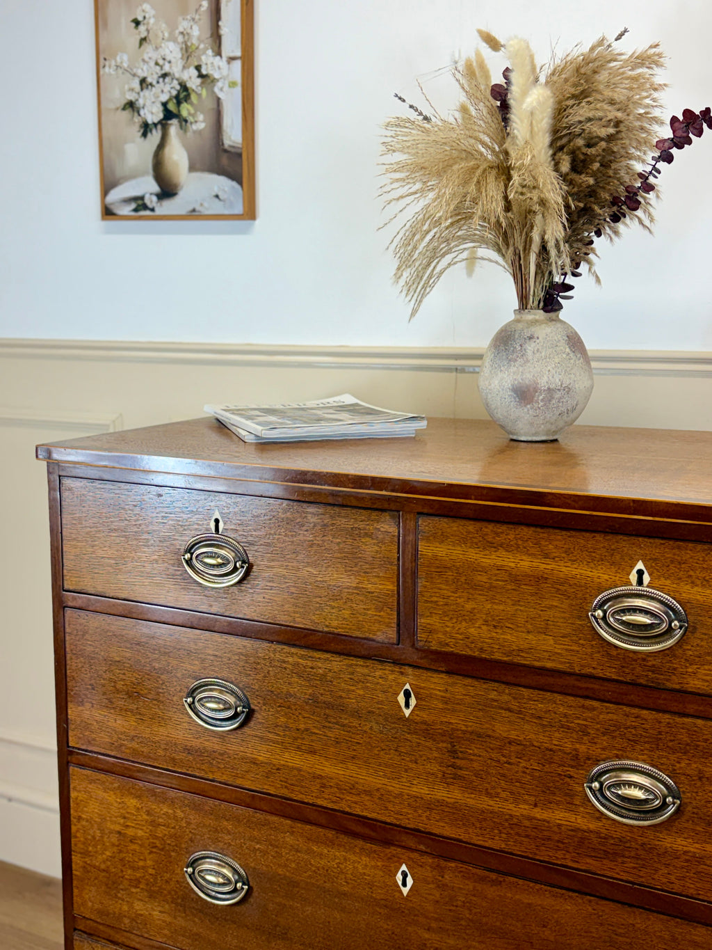 Wooden dresser with decorative items including a vase with dried plants and a framed picture on a wall.
