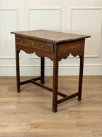 Wooden side table with a single drawer on a wooden floor against a white paneled wall.