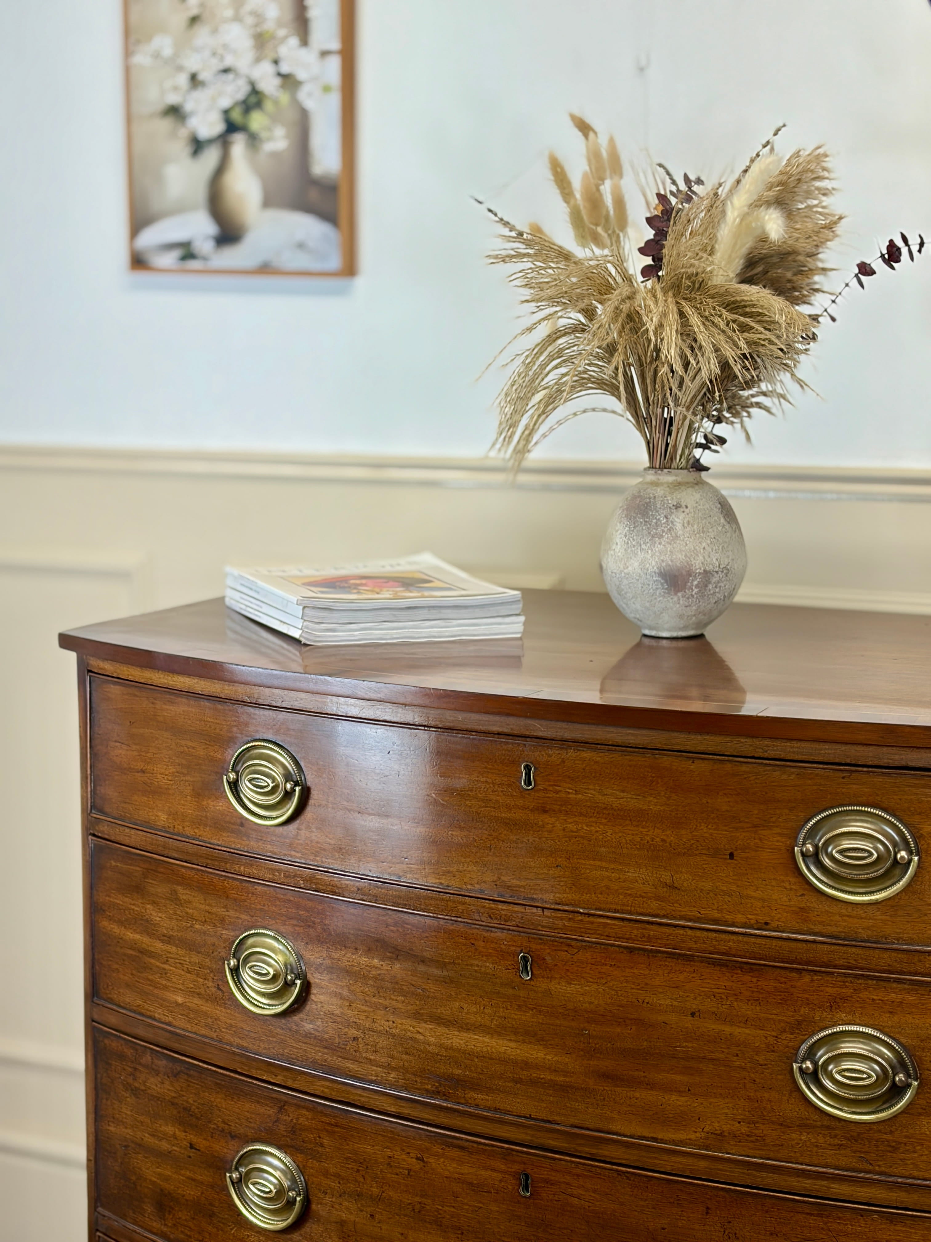 Wooden dresser with brass handles featuring a vase of dried plants and books on top.