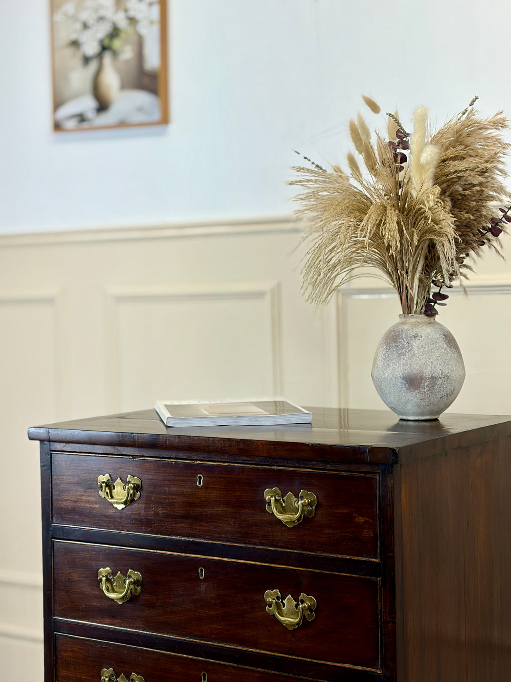 Wooden dresser with a vase of dried plants and a tablet on top, against a light wall.