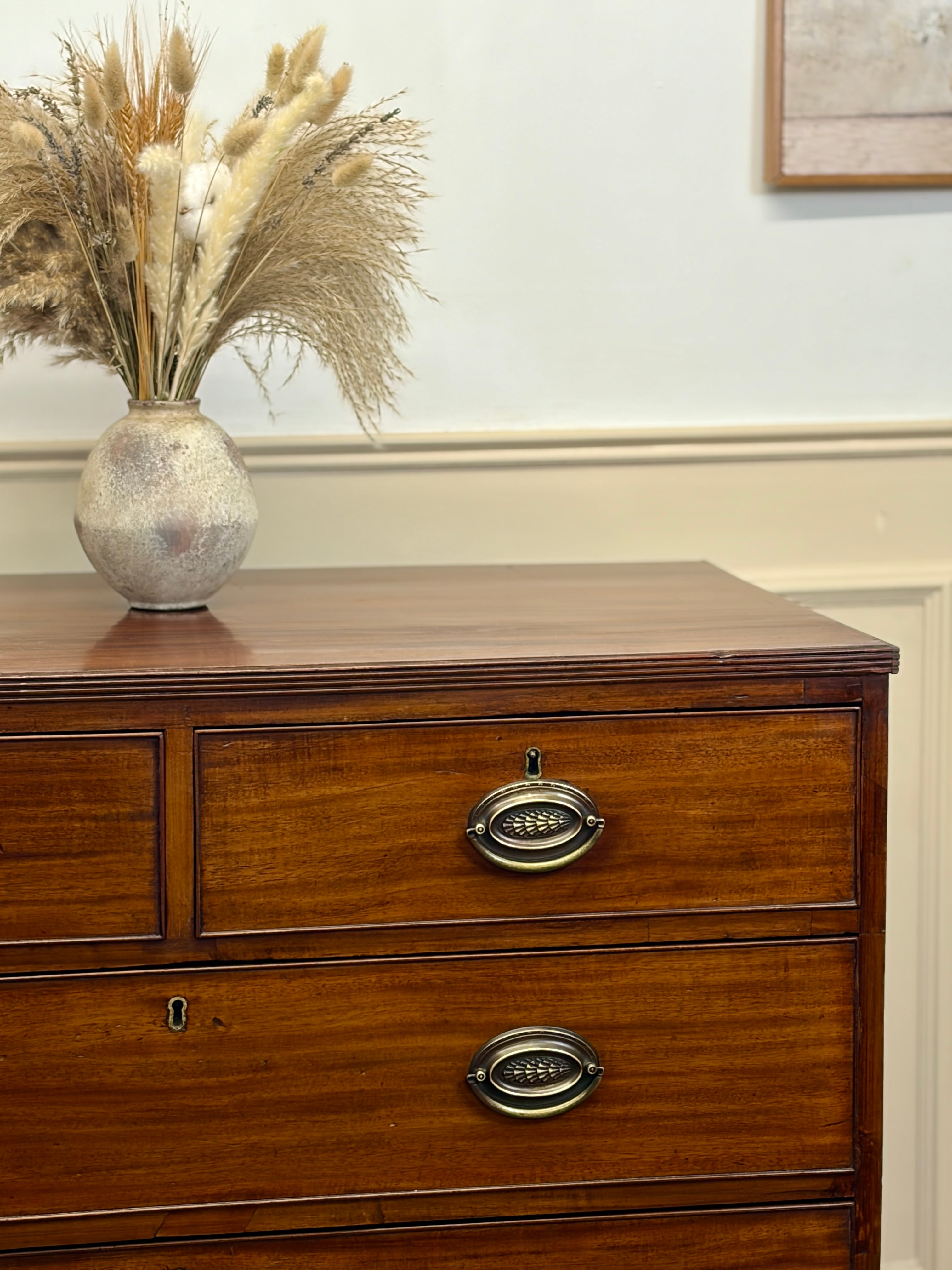 Wooden dresser with decorative vase and dried plants on top against a white wall.
