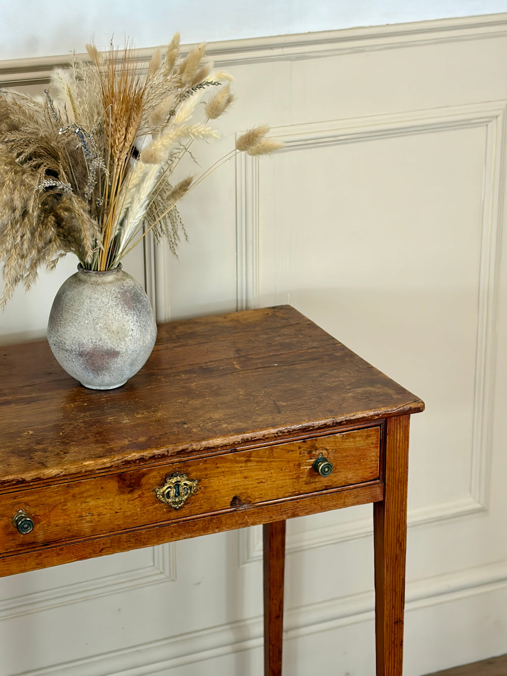 Vintage wooden side table with a vase of dried pampas grass against a white paneled wall.