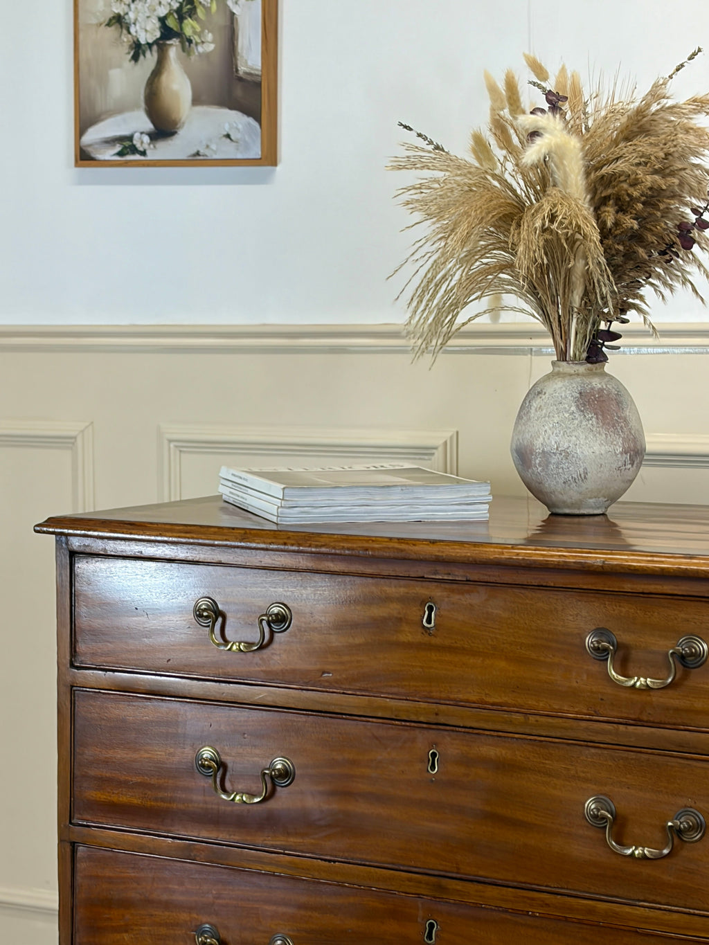 Wooden dresser with a vase of dried plants and books on top, framed picture in the background.