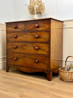 Wooden dresser with four drawers on a wooden floor, with a basket and decorative vase in the background.