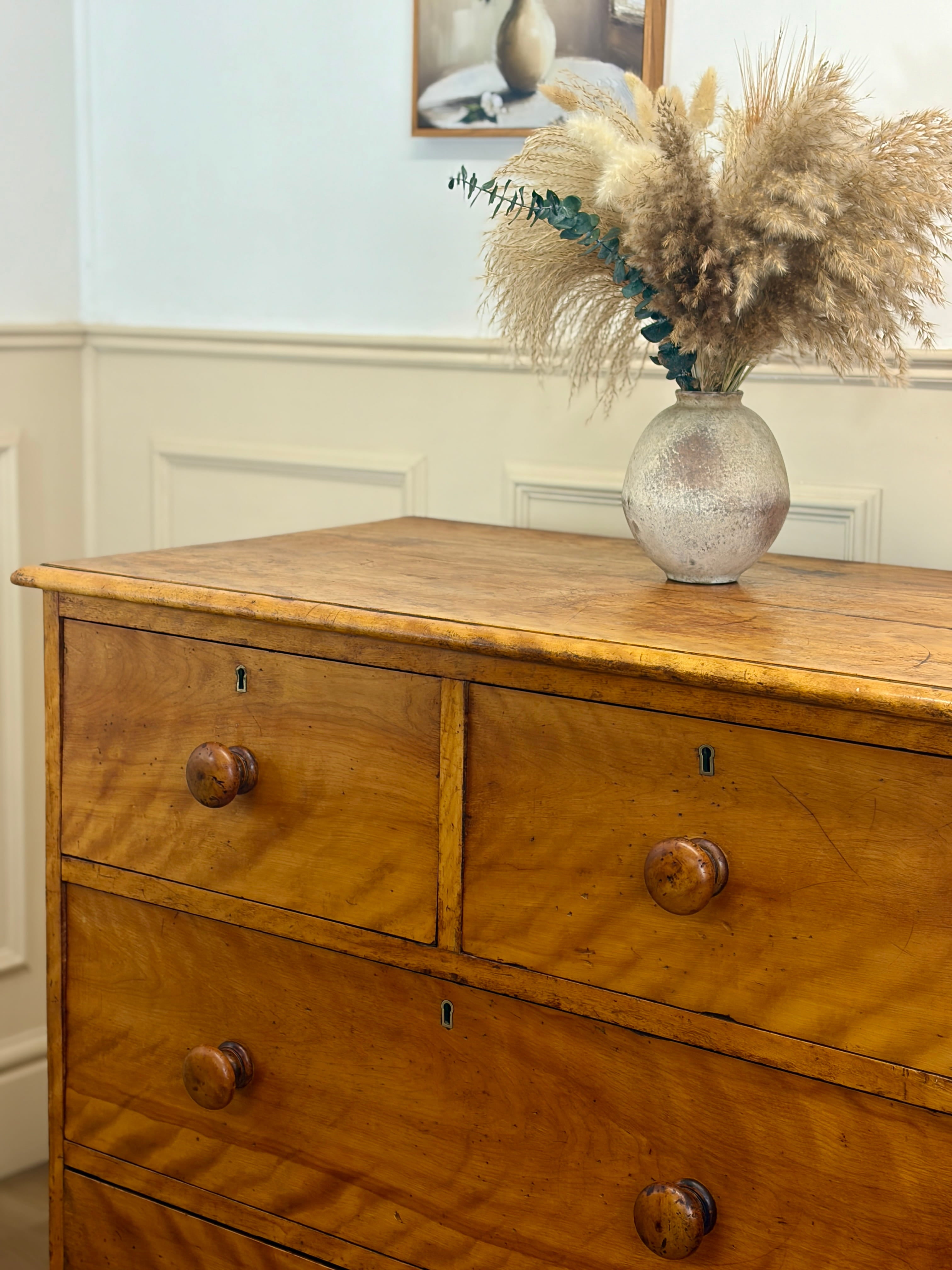 Wooden dresser with a vase of dried plants on top, against a light-colored wall.