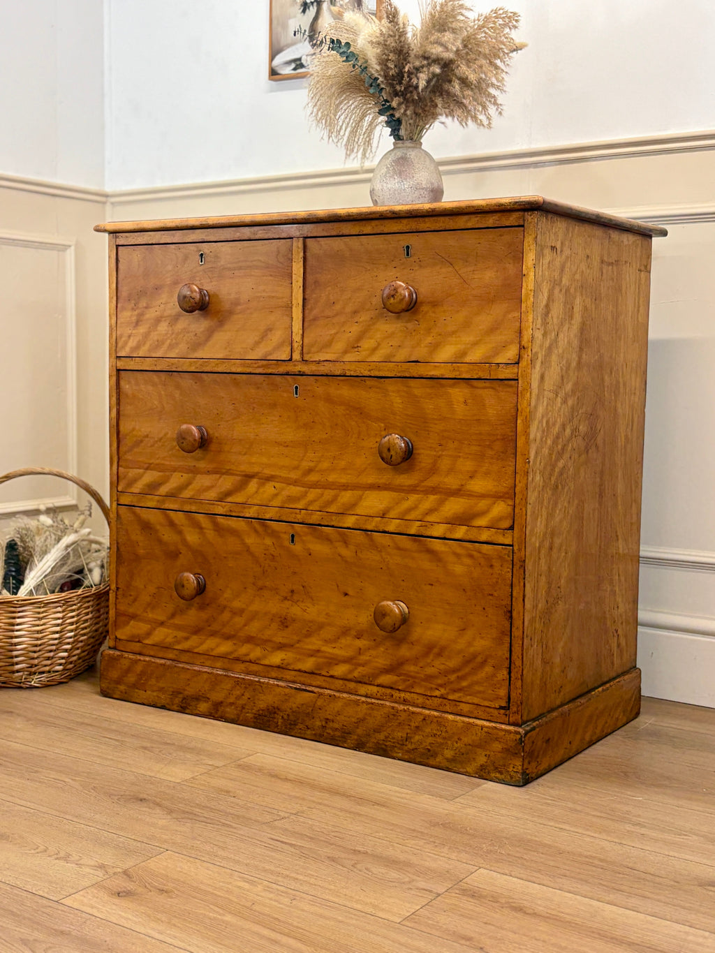 Wooden dresser with three drawers on a wooden floor, next to a basket and a vase with dried plants.
