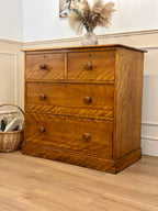 Wooden dresser with three drawers on a wooden floor, next to a basket and a vase with dried plants.