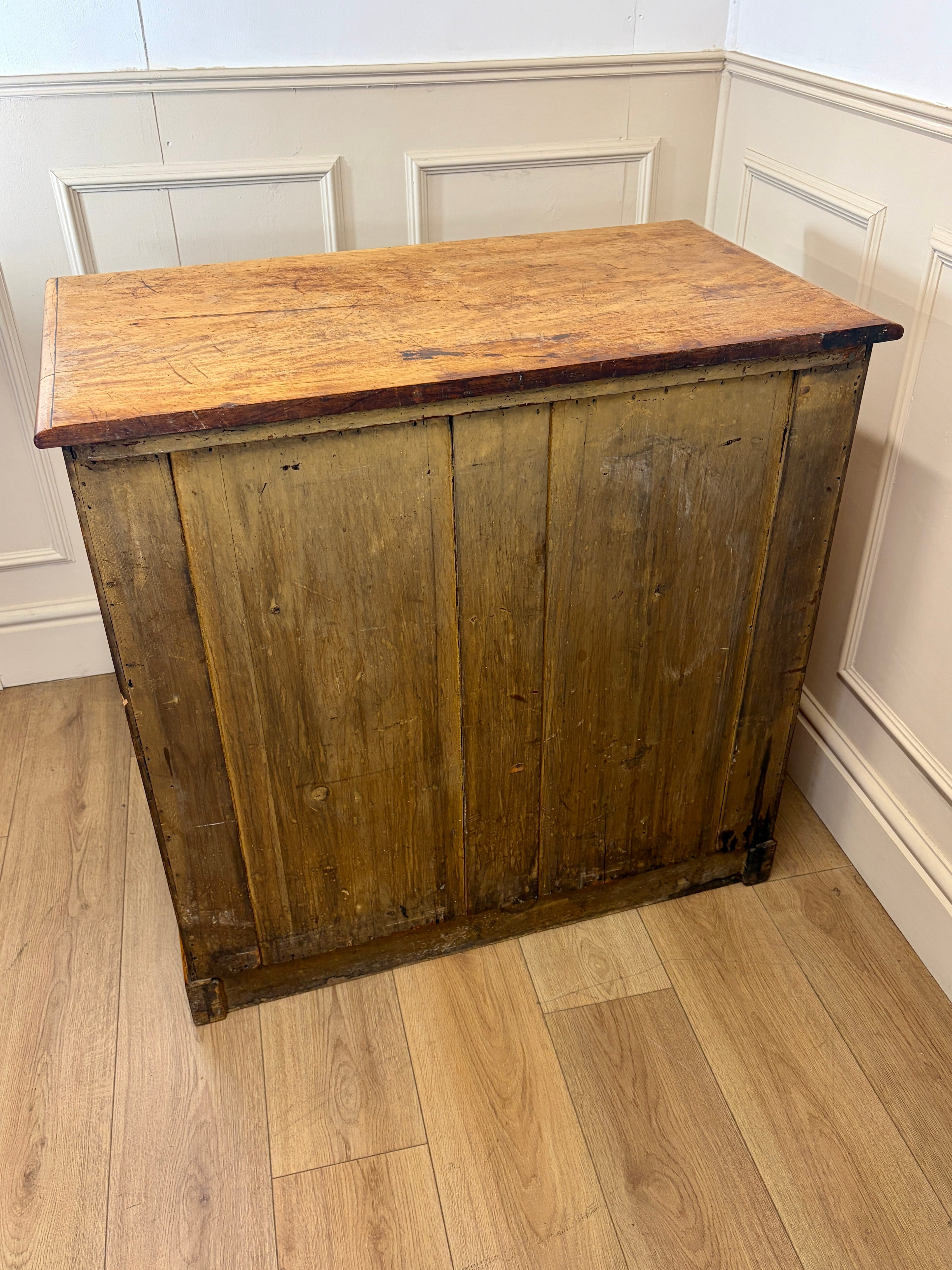 Wooden cabinet with a worn finish on a wooden floor against a white paneled wall.