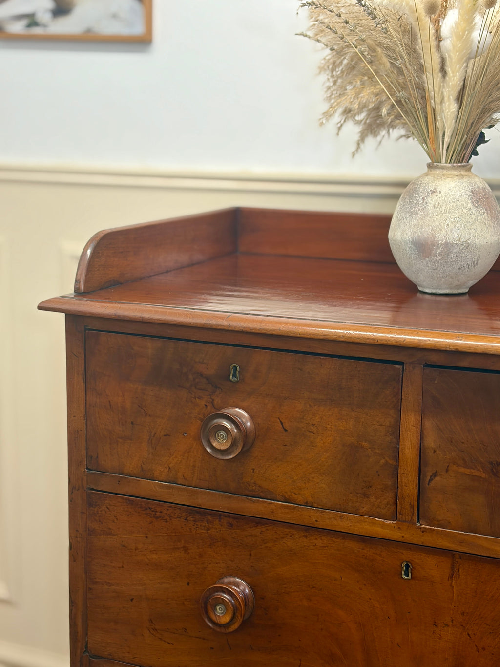 Wooden dresser with a vase of dried plants on top against a white wall.