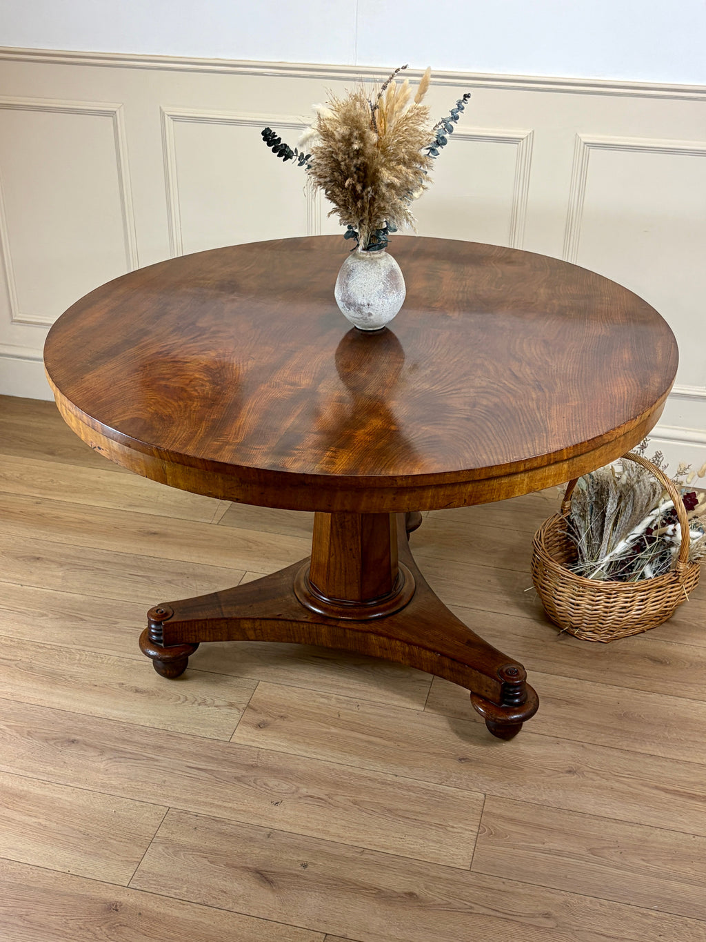 Round wooden table with a vase of dried flowers on a wooden floor.