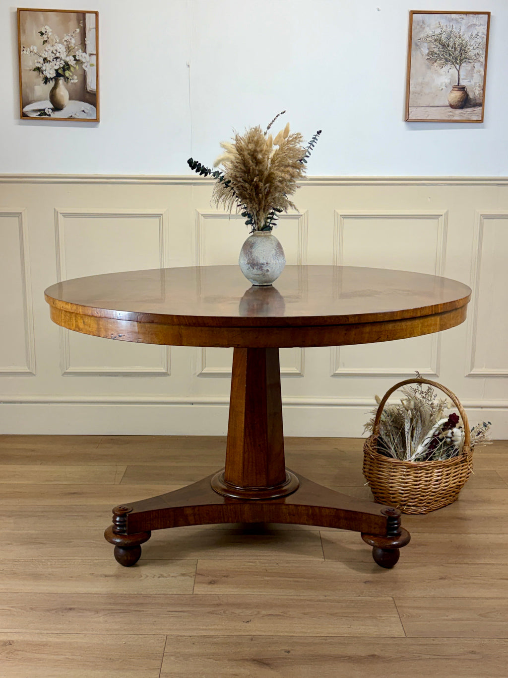 Wooden round table with a vase of flowers on a wooden floor, with two framed pictures on the wall.