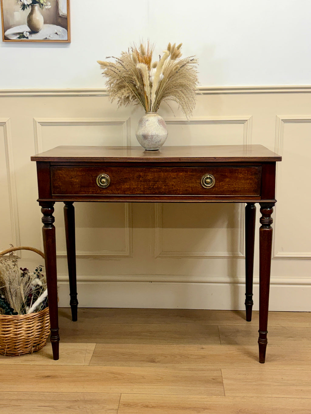 Wooden console table with a vase of dried plants against a white paneled wall.