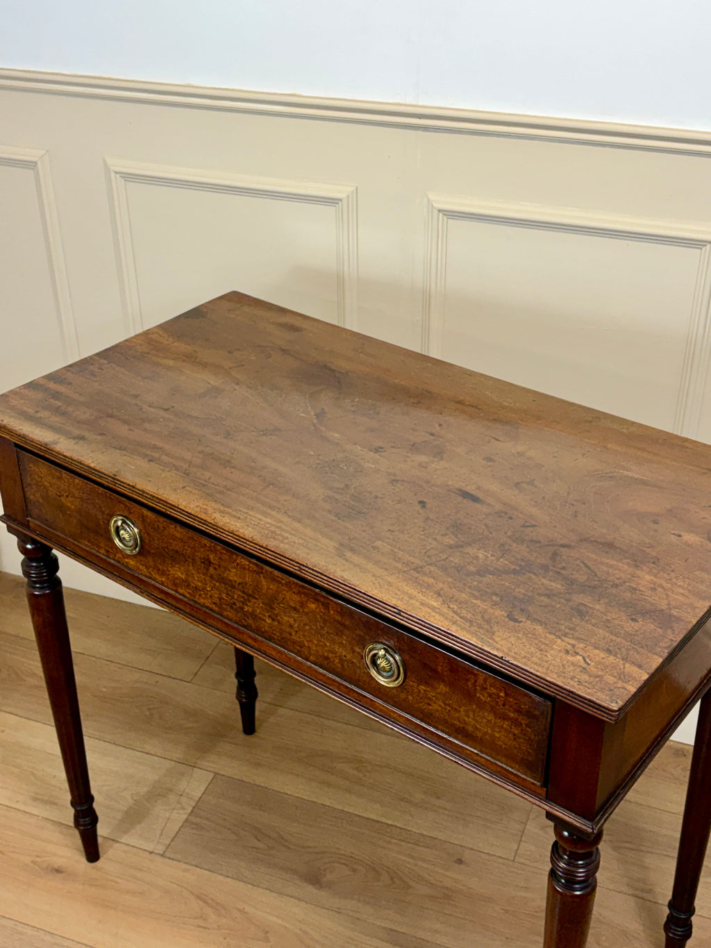 Wooden desk with a single drawer against a white paneled wall.
