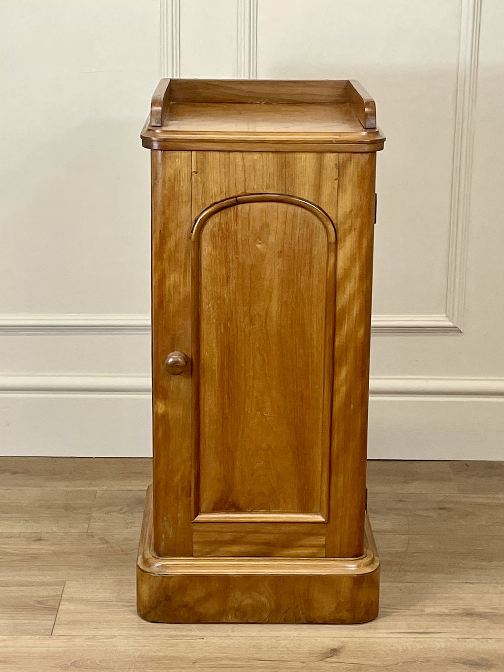 Wooden cabinet with a single door on a wooden floor against a white paneled wall.