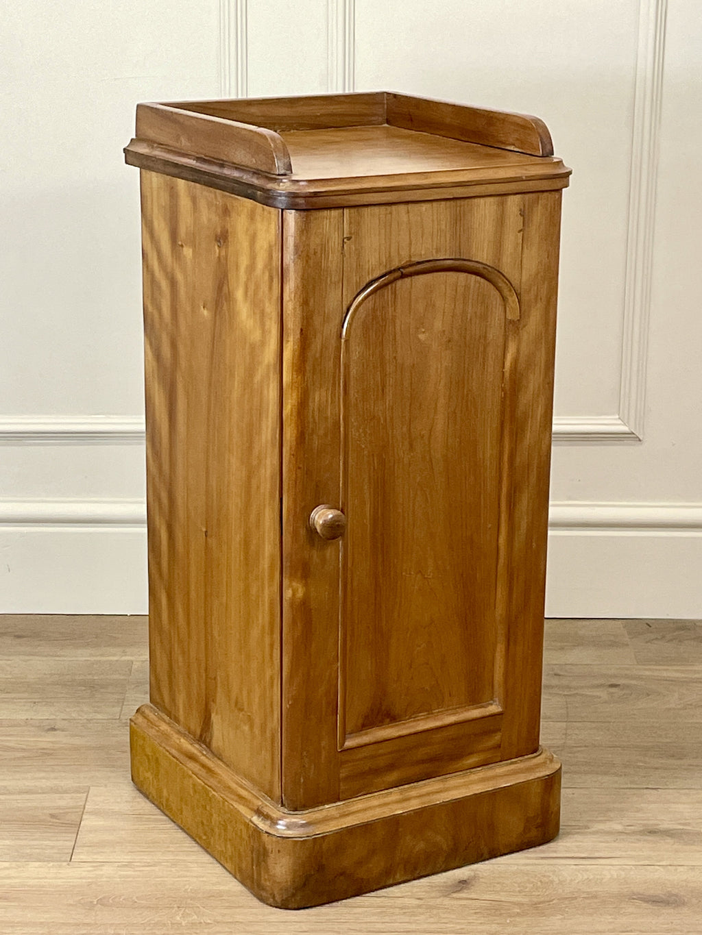 Wooden cabinet with a single door on a wooden floor against a white paneled wall.