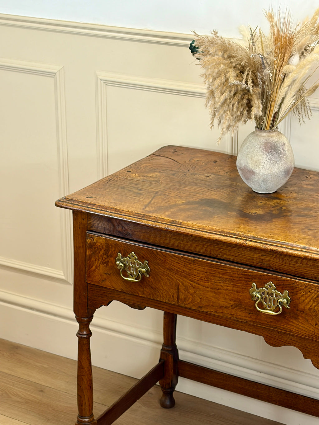Wooden side table with a vase of dried flowers against a white paneled wall.