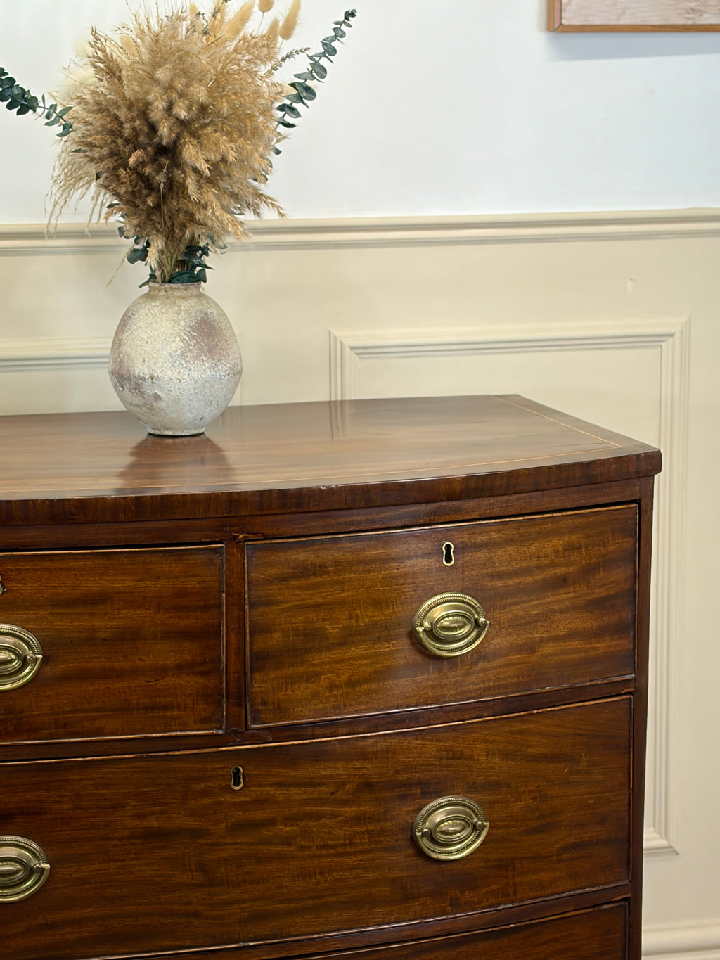 Wooden dresser with brass handles and a vase with dried plants on top against a white wall.