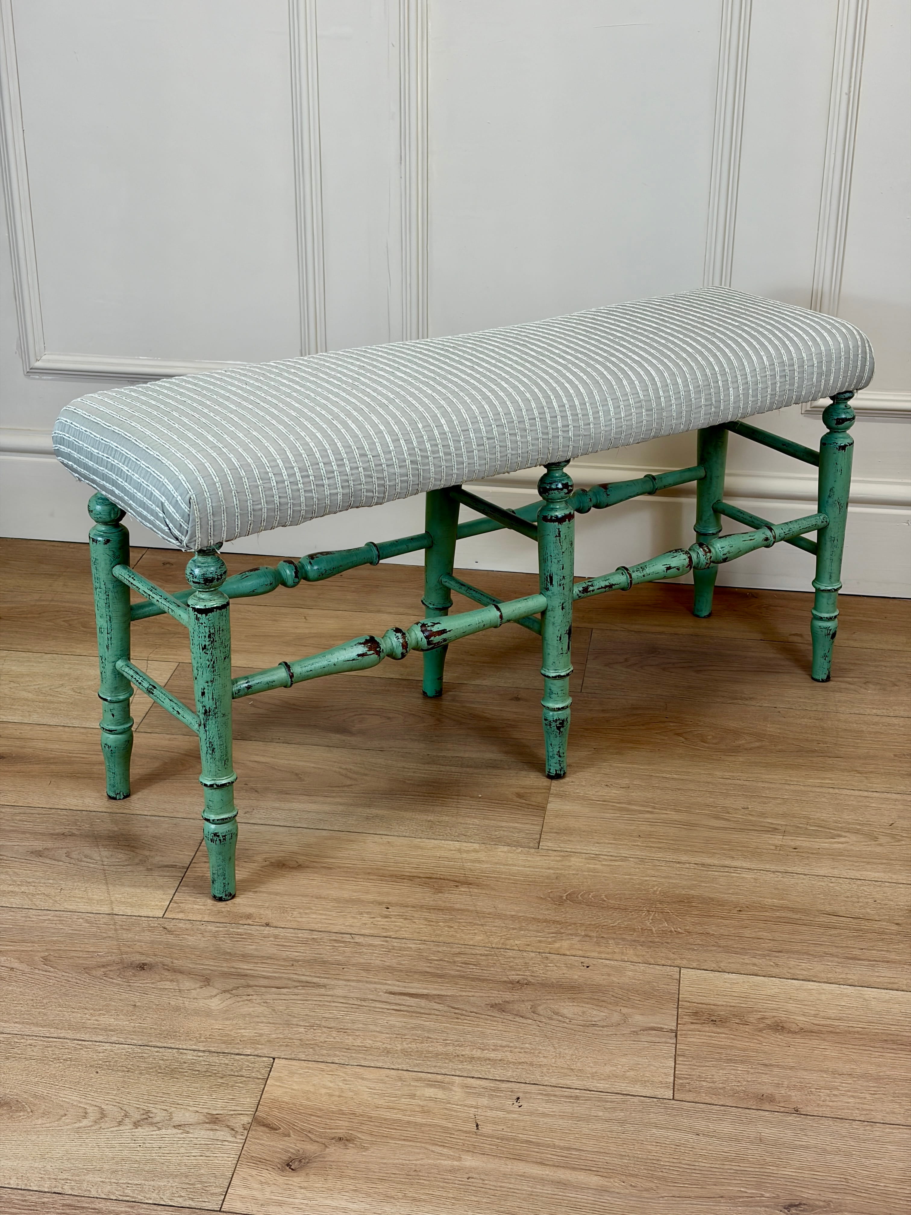 Green bench with striped cushion on a wooden floor against a white paneled wall.
