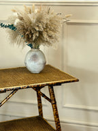 Decorative vase with dried plants on a wicker table against a neutral wall.