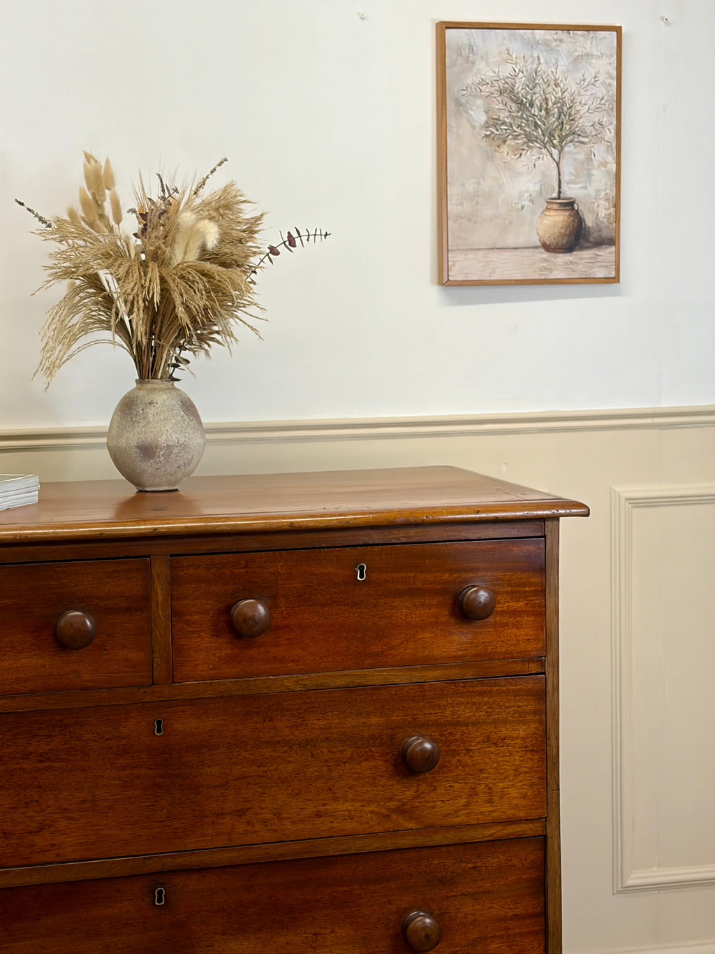 Wooden dresser with a vase of dried plants and a framed picture on the wall.