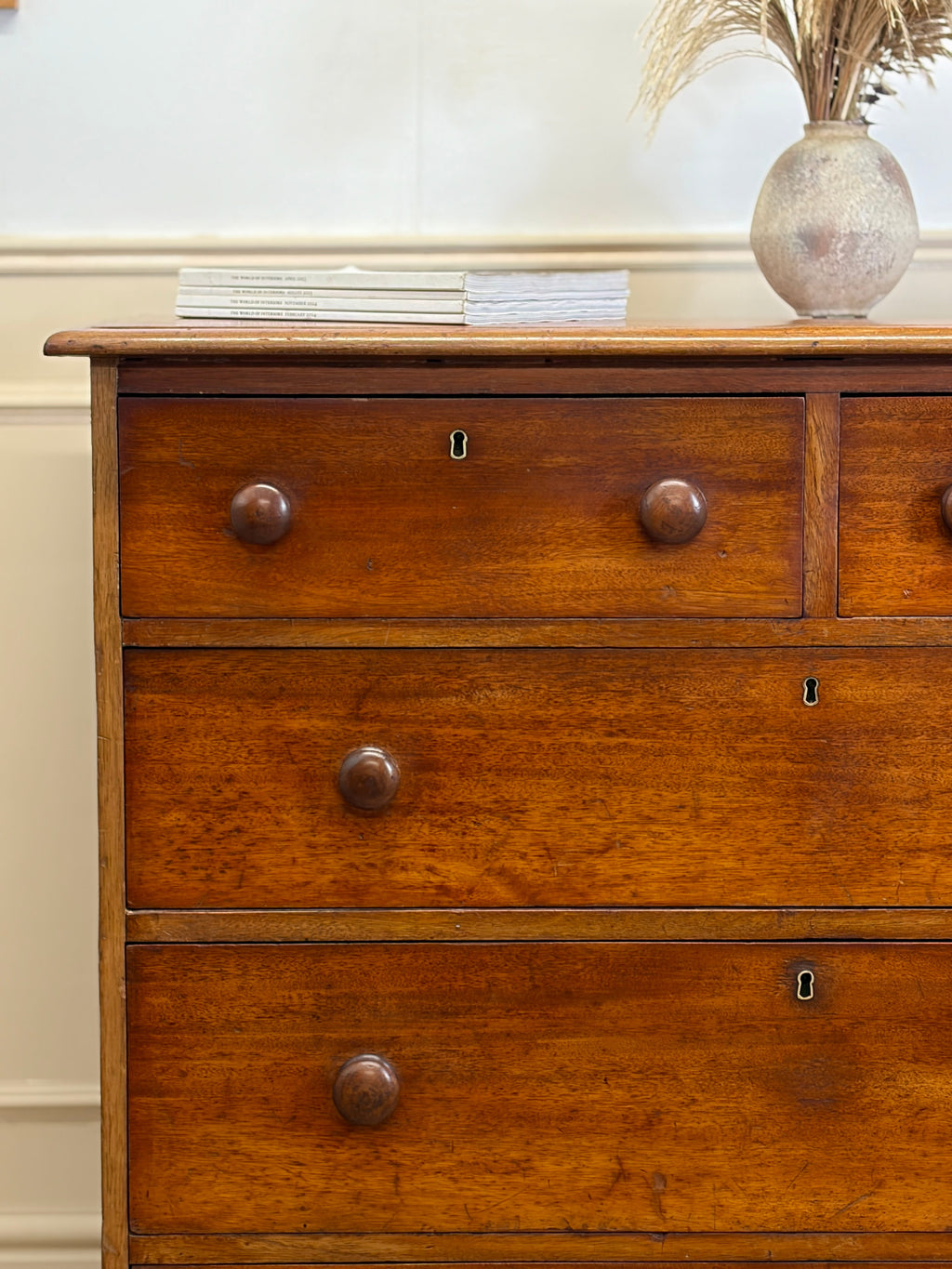 Wooden dresser with three drawers and a vase on top against a neutral wall.