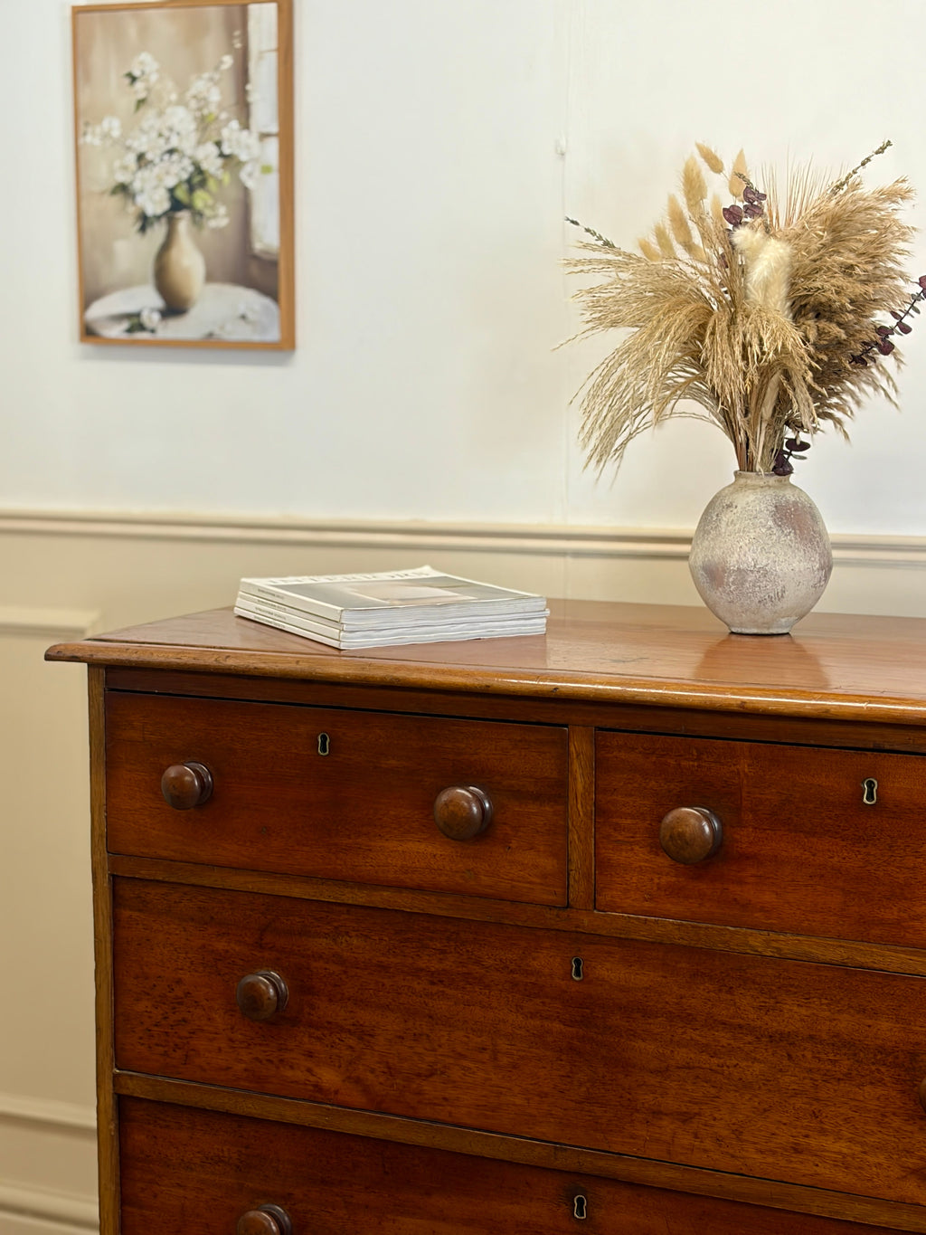Wooden dresser with a vase of flowers and books on top, against a white wall.