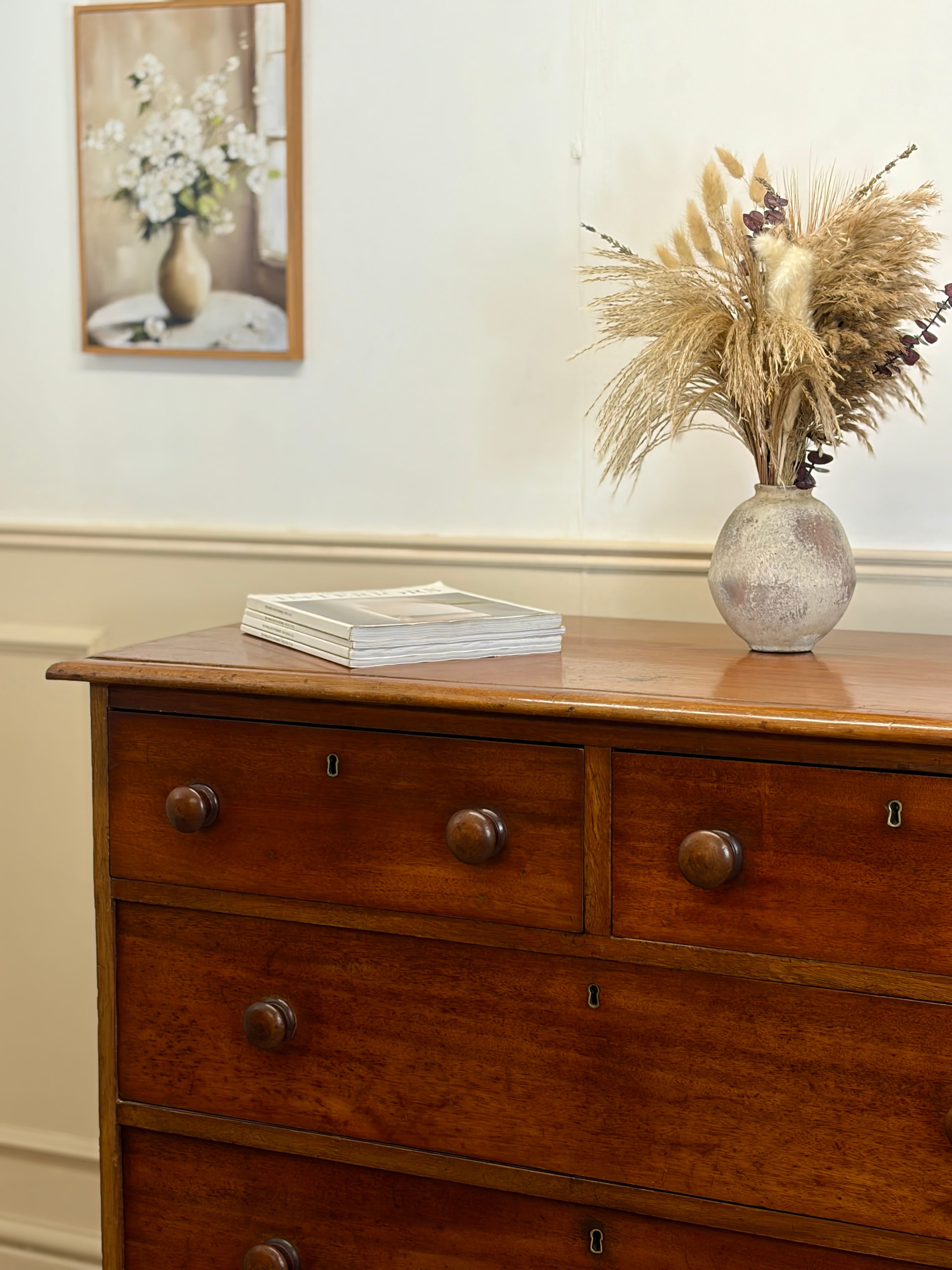 Wooden dresser with a vase of flowers and books on top, against a white wall.