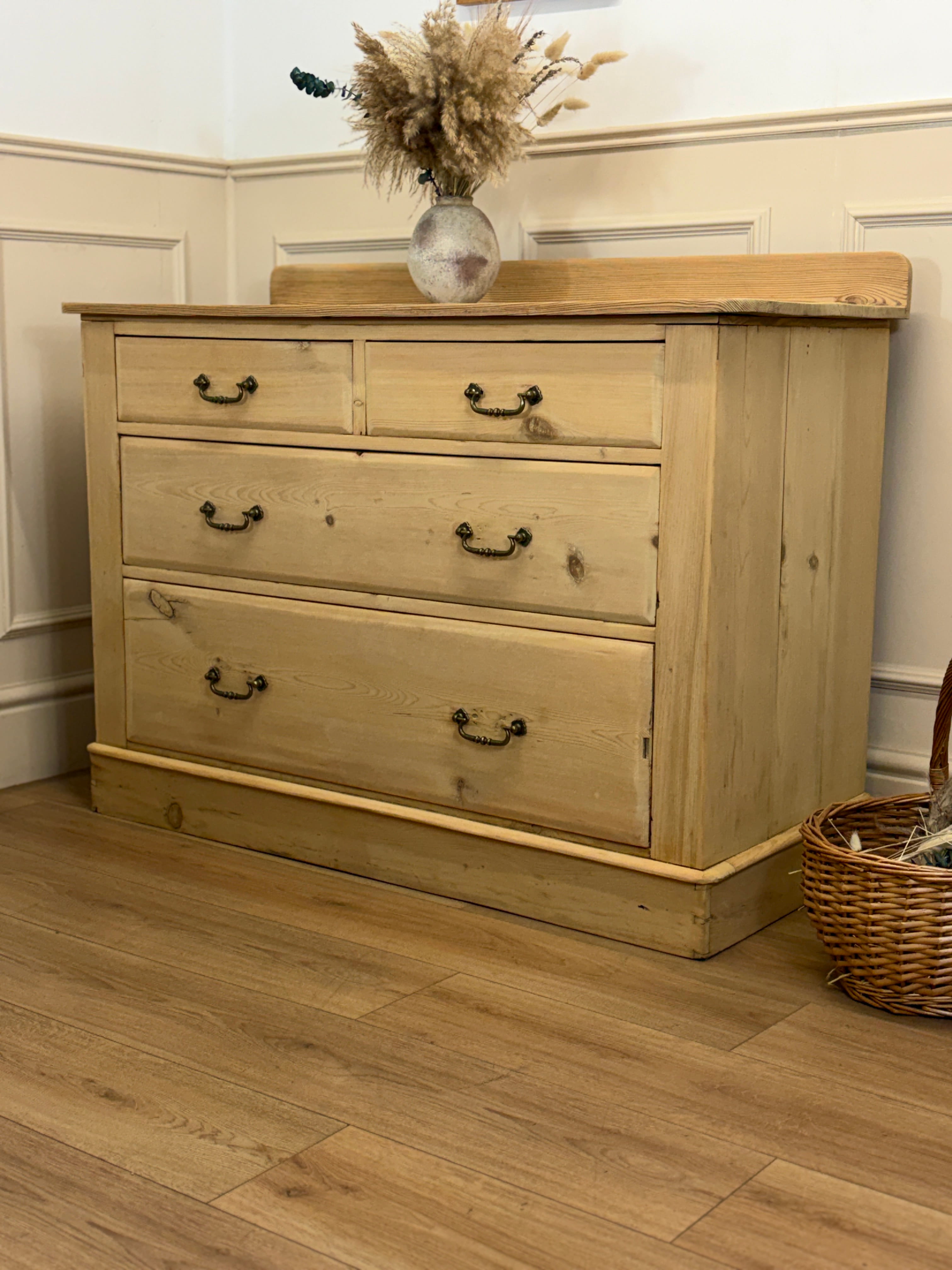 Wooden dresser with three drawers on a wooden floor, featuring a vase with dried flowers on top.