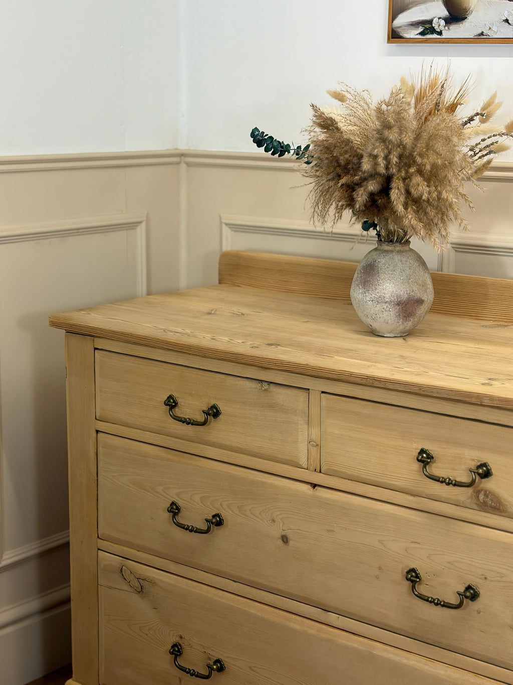Wooden dresser with a vase of dried flowers on top against a white wall.