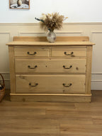 Wooden dresser with four drawers and a vase of flowers on top in a room with white walls and wooden floor.