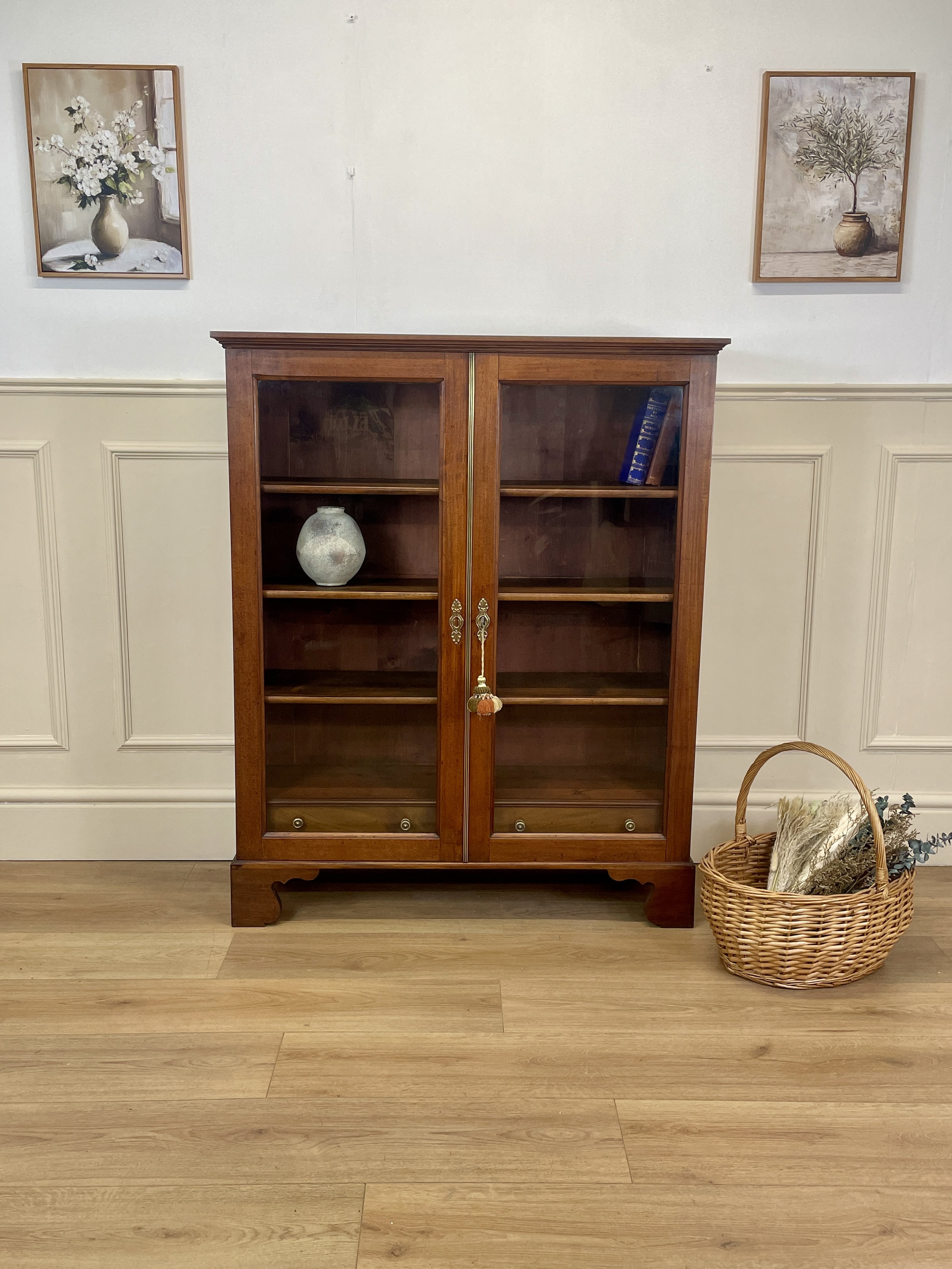 Wooden bookshelf with glass doors in a room with wainscoting and framed pictures.