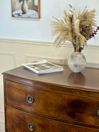 Wooden dresser with a vase of dried plants and a newspaper on top, against a light wall.