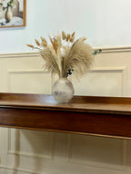 Decorative vase with dried plants on a wooden surface against a light wall.