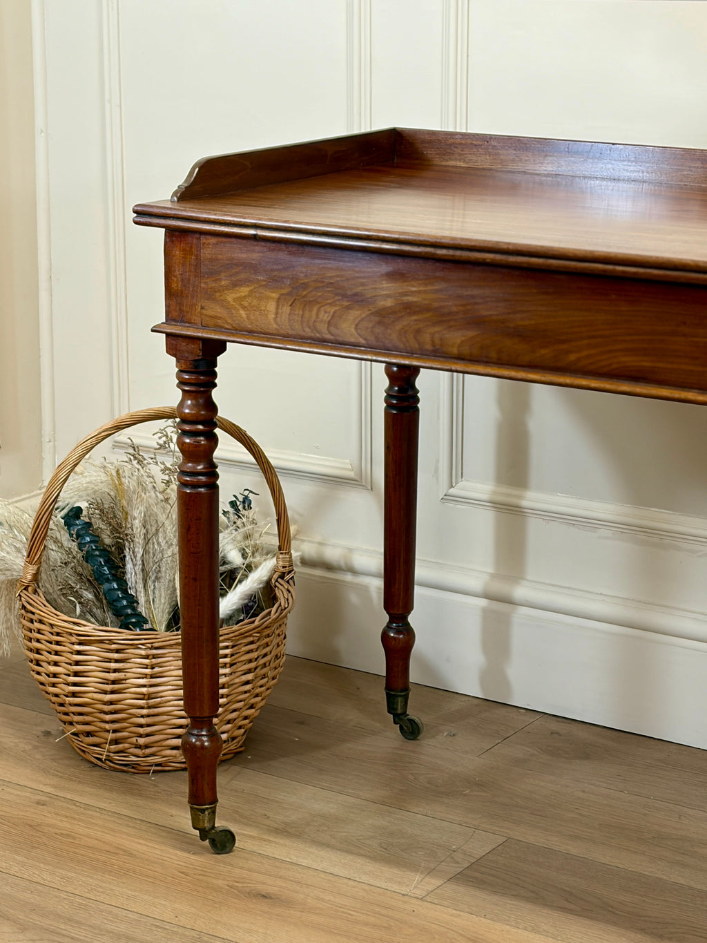 Wooden side table with a wicker basket on a wooden floor against a white paneled wall.