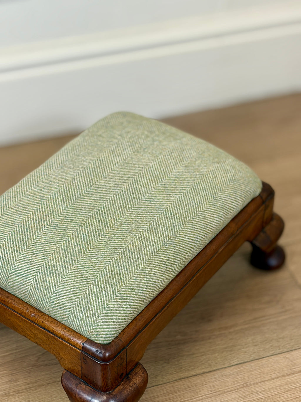 A small wooden stool with a light sage green herringbone upholstered cushion, shown on a light wood floor against a white wall.
