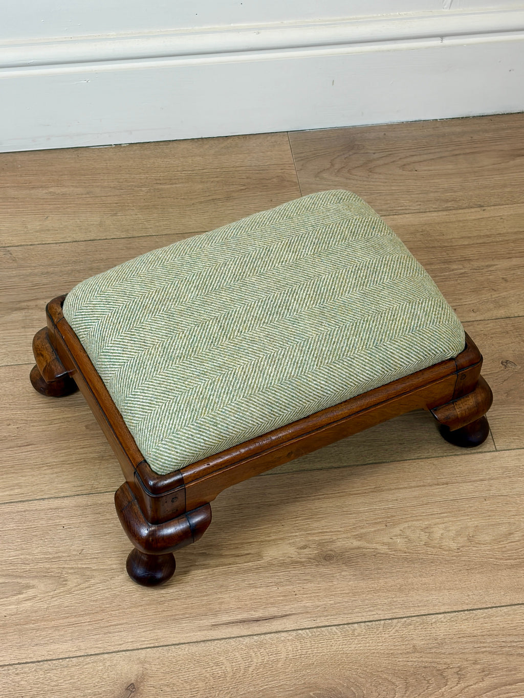 A small wooden stool with a square light green herringbone upholstered cushion, set on a light wood floor against a white baseboard.