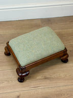 A small wooden stool with a square light green herringbone upholstered cushion, set on a light wood floor against a white baseboard.