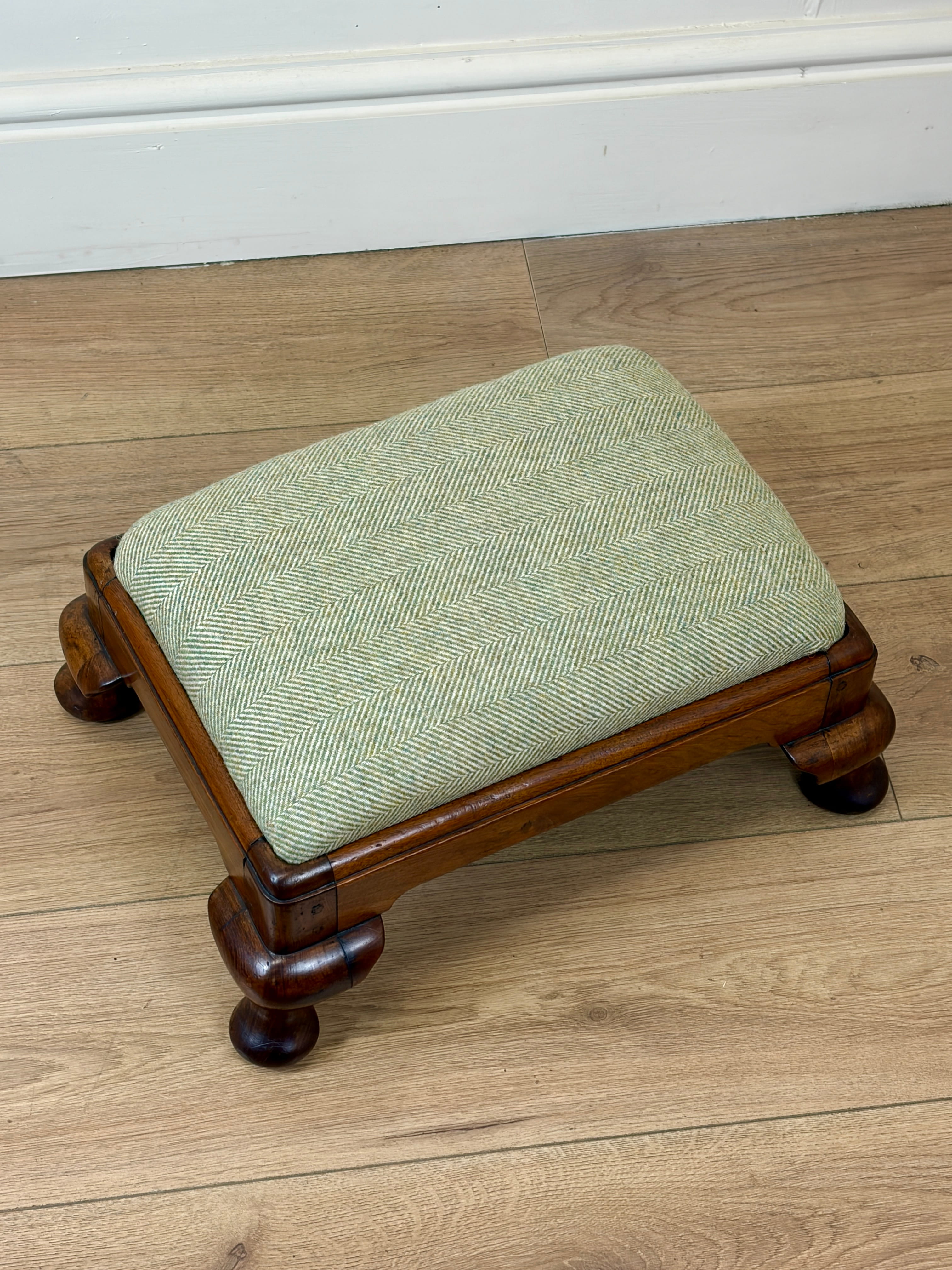 A small wooden stool with a square light green herringbone upholstered cushion, set on a light wood floor against a white baseboard.