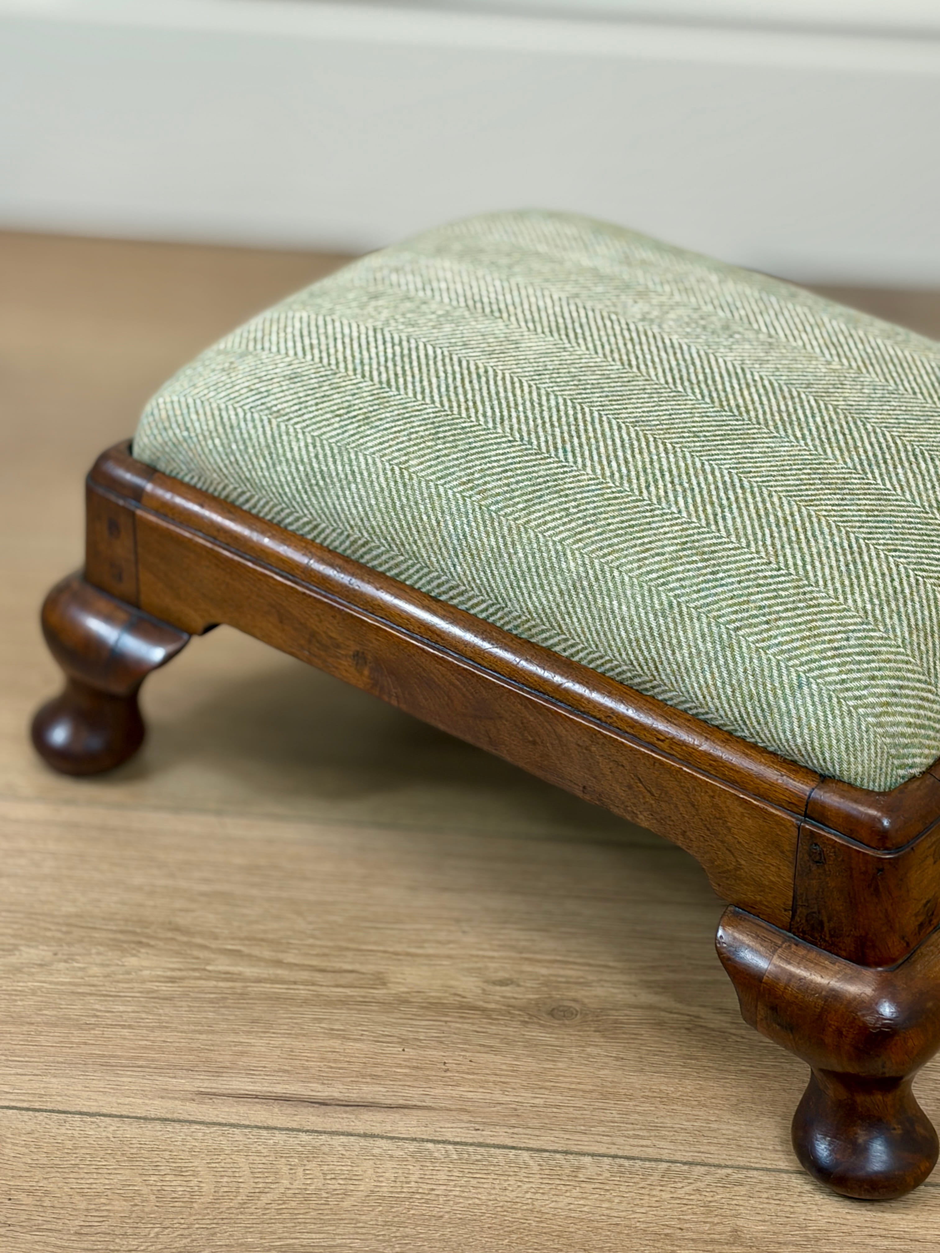 A small wooden stool with a light sage green herringbone upholstered cushion, shown on a light wood floor against a neutral wall.