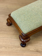 A close-up of an upholstered bench or stool with a light sage green and beige herringbone fabric cushion and a dark brown wooden frame with turned legs, shown on a light wood floor.