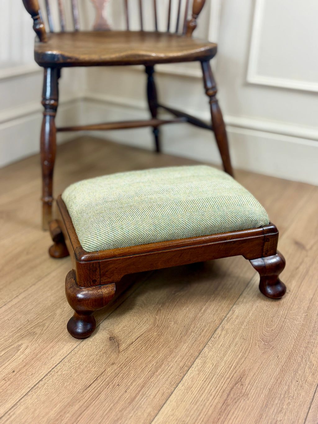 A small wooden footstool with a light sage green herringbone upholstered cushion, positioned in front of a dark wood spindleback chair on a light oak floor.