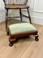 A small wooden footstool with a light sage green herringbone upholstered cushion, positioned in front of a dark wood spindleback chair on a light oak floor.