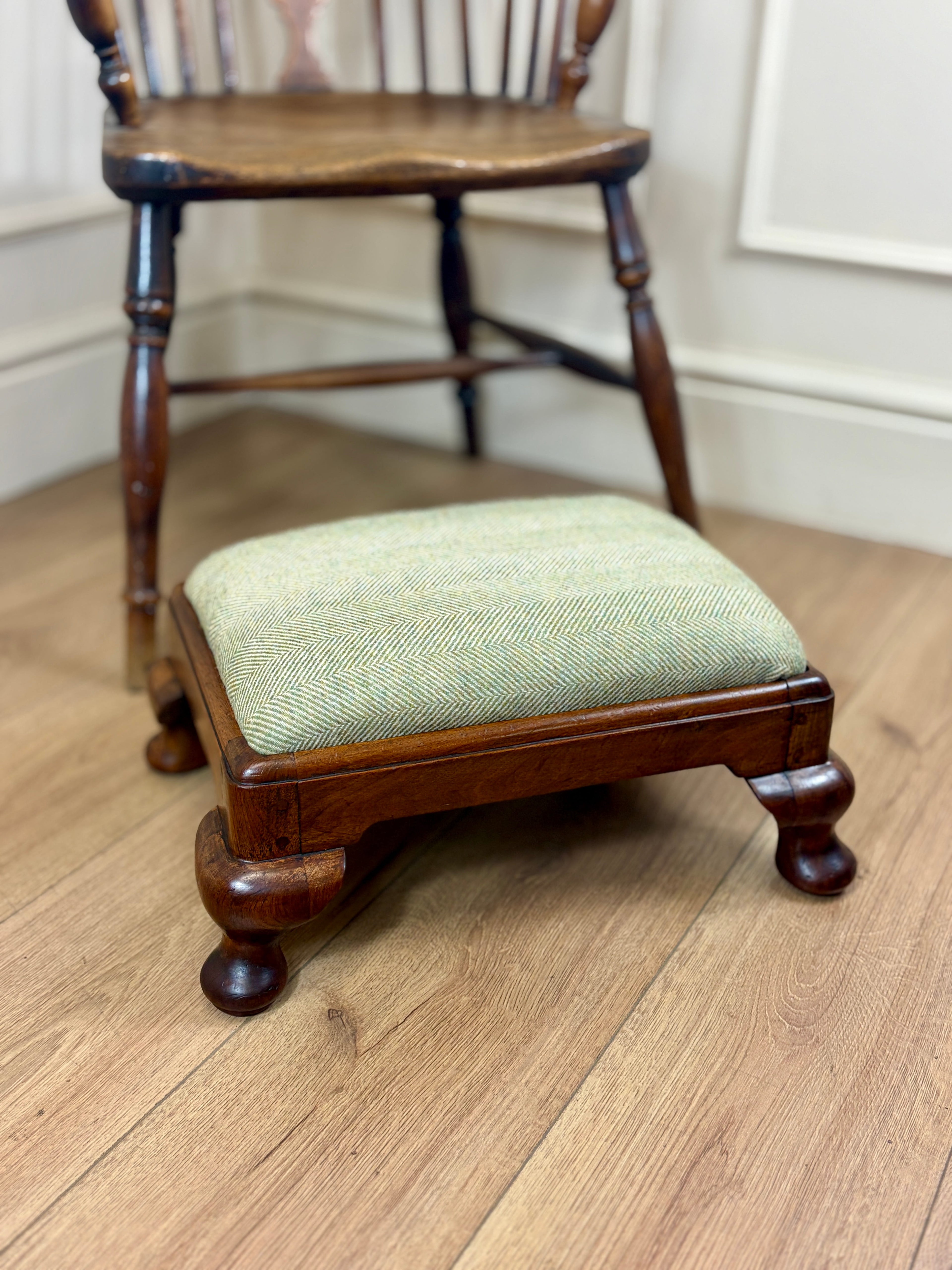 A small wooden footstool with a light sage green herringbone upholstered cushion, positioned in front of a dark wood spindleback chair on a light oak floor.