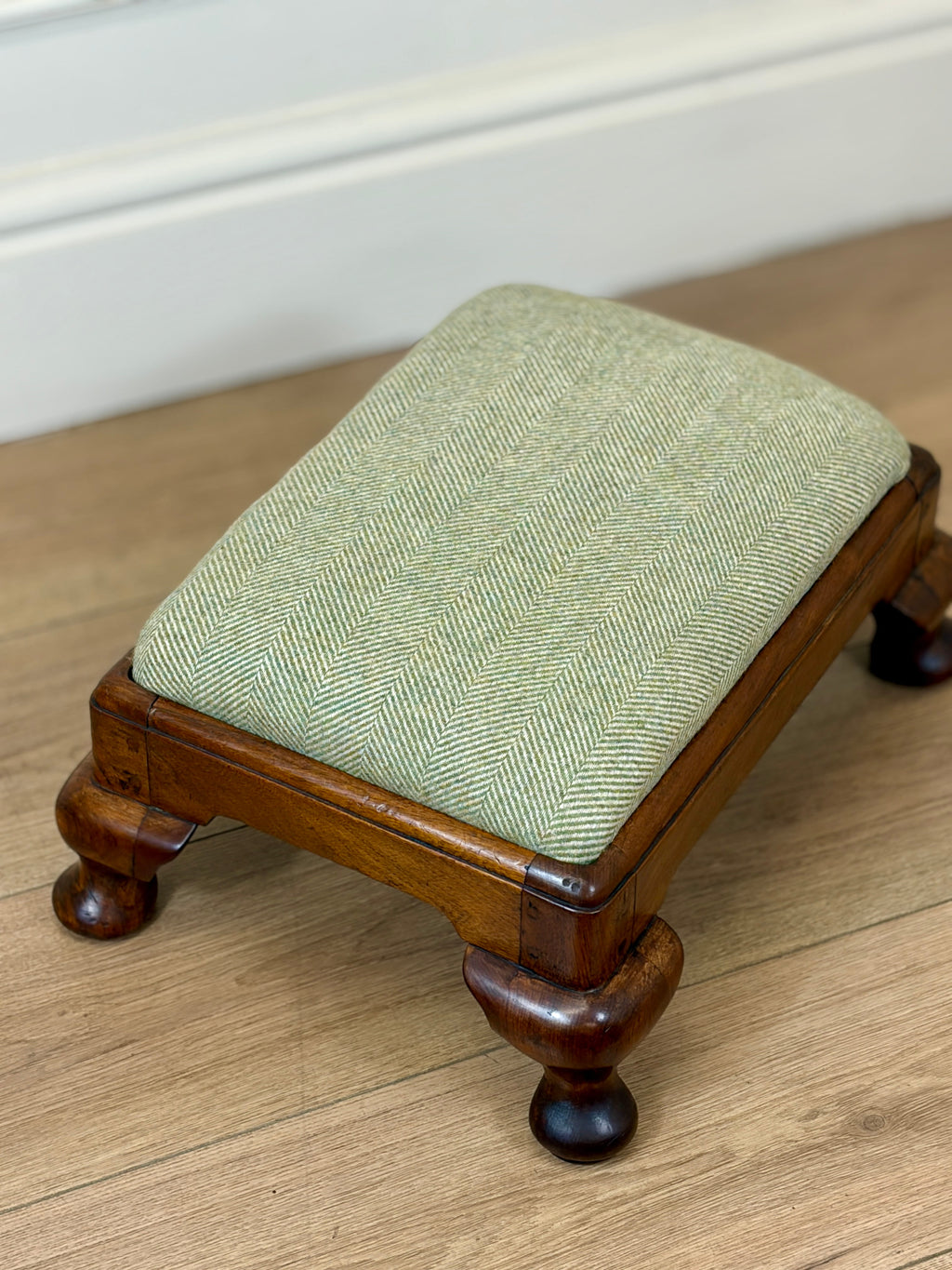 A small wooden footstool with a light sage green herringbone upholstered cushion, shown on a light wood floor against a white wall.